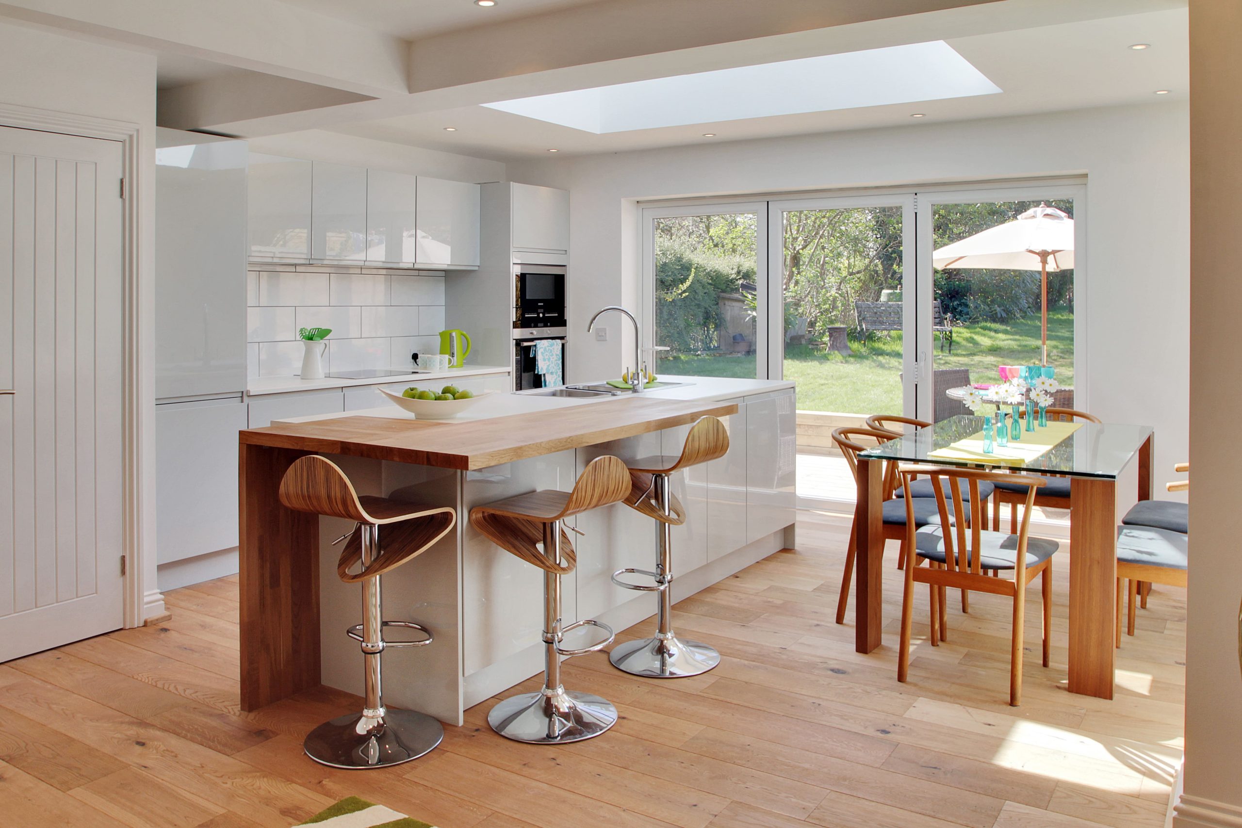 Gloss white handleless kitchen with oak breakfast bar island, timber bar stools, and bifold doors opening to garden, by Jones Britain Tunbridge Wells