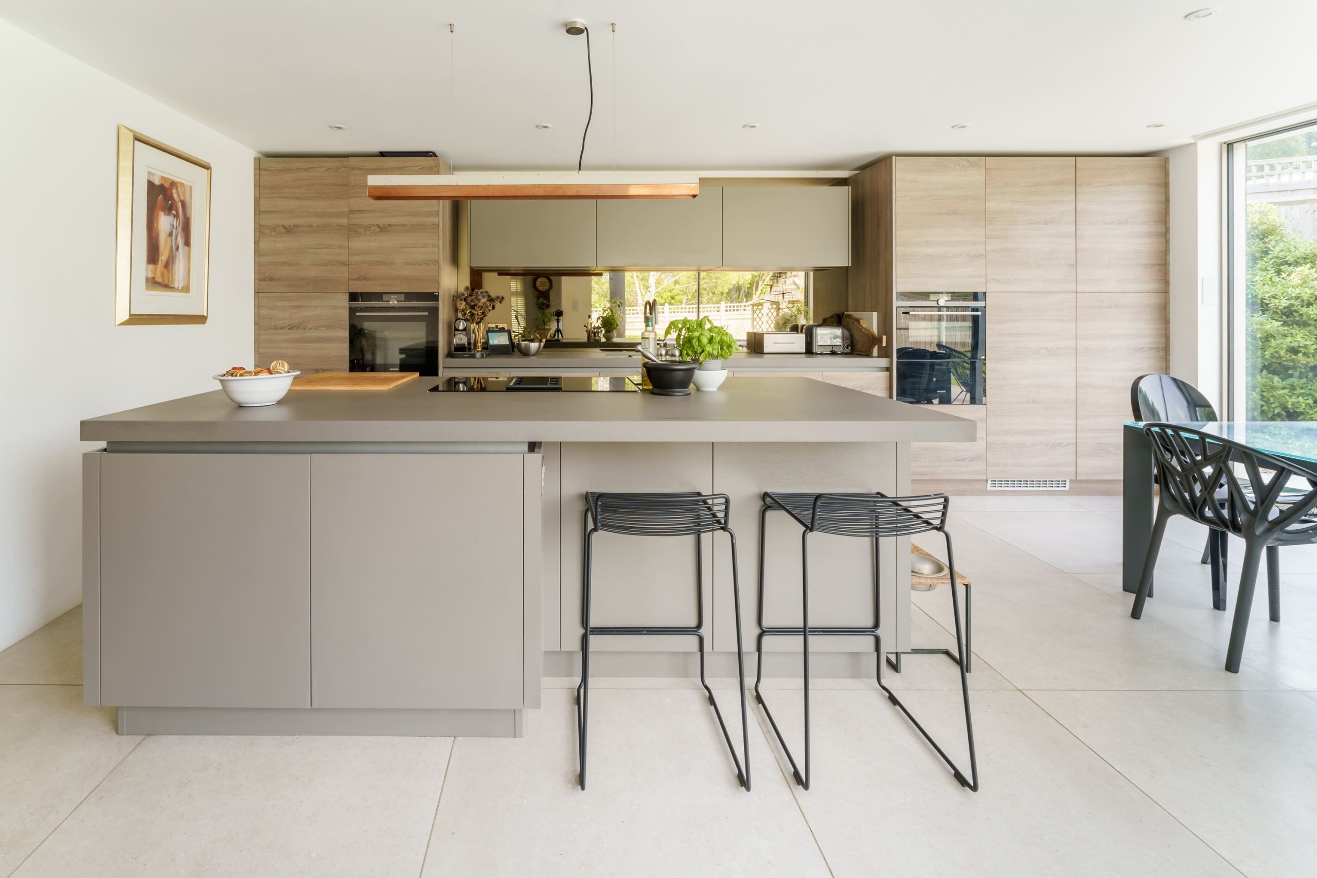 Greige handleless kitchen with stone worktop island, light oak tall units, mirrored splashback, and open-plan dining area, designed by Jones Britain