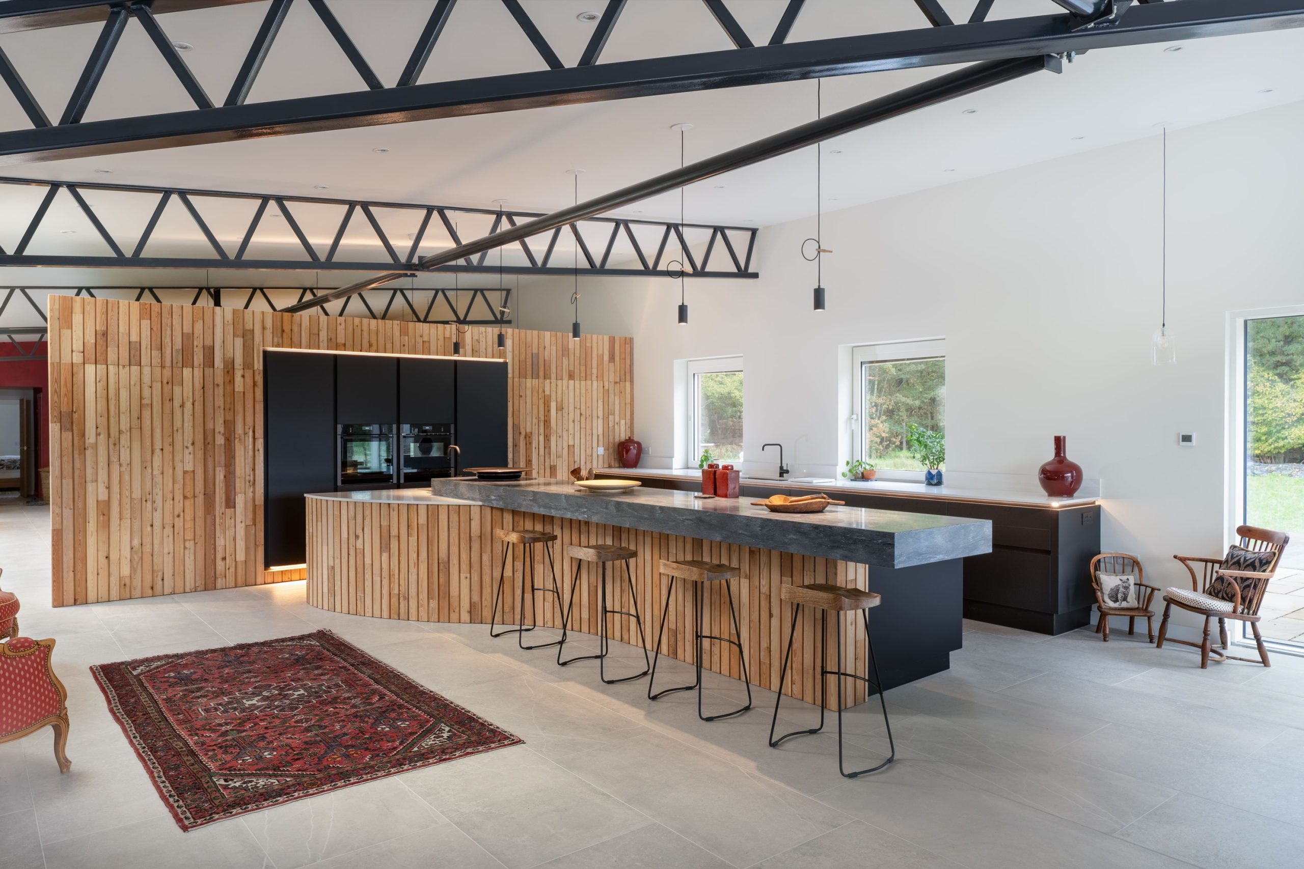 Bespoke open-plan handleless kitchen with timber clad island, dark stone worktop, black pendant lighting, and Persian rug, by Jones Britain Tunbridge Wells