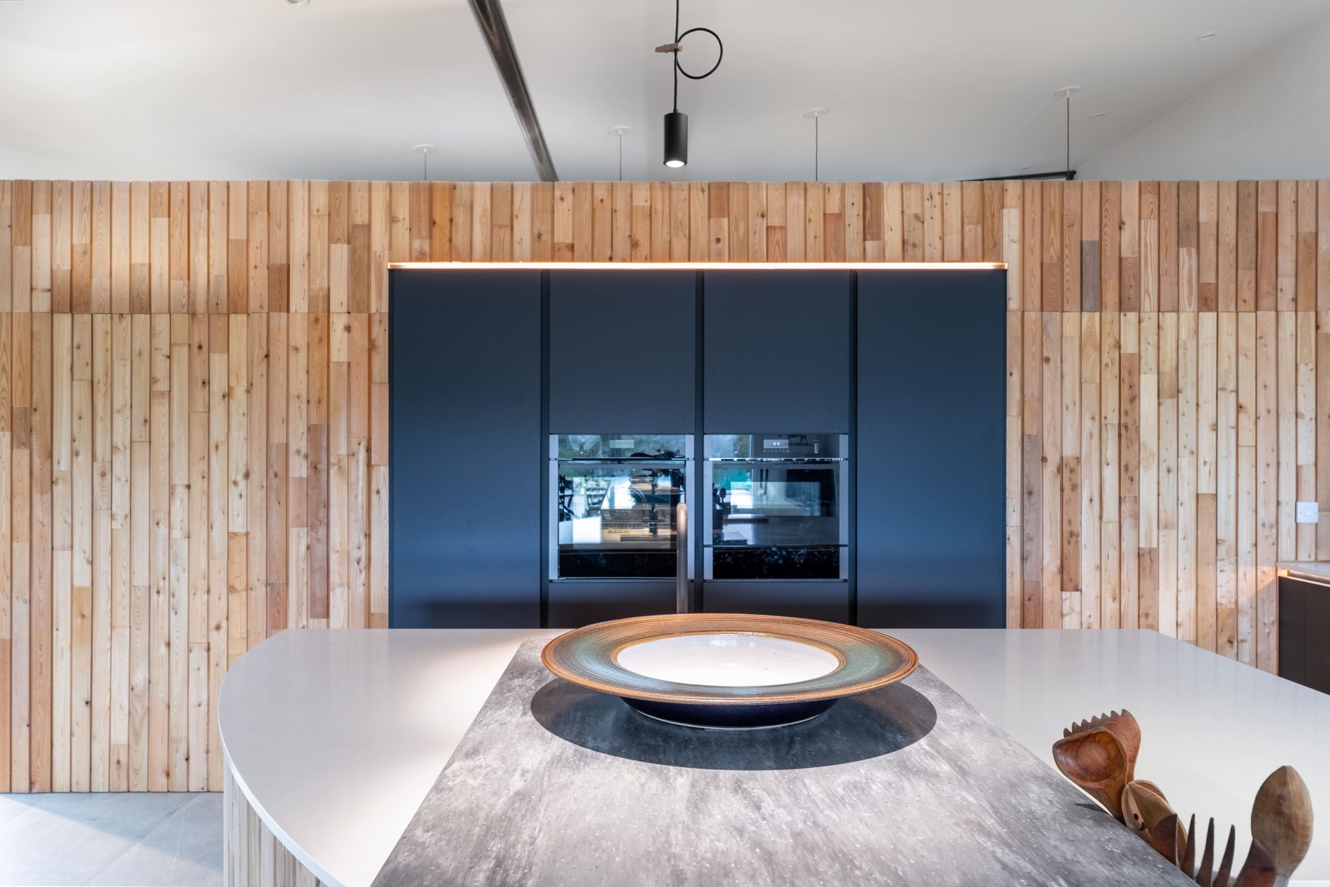 Bespoke black handleless tall units with integrated ovens set into a vertical timber clad feature wall, with curved stone island, by Jones Britain